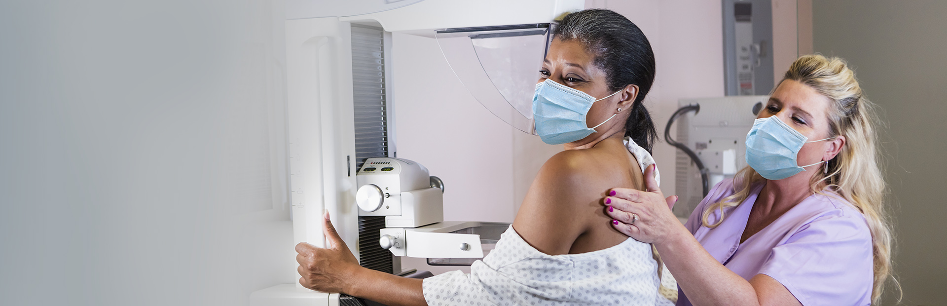 一名40多岁的成熟非裔美国妇女穿着医院的大褂，接受每年一次的乳房x光检查，在一名技术人员的帮助下，一个穿着手术服的金发女人。